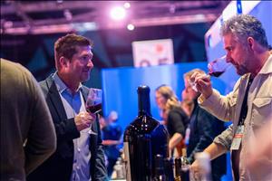 Two men looking at each other as they hold glasses of wine in front of other vendor stalls